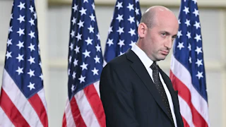 Stephen Miller glares as he stands in front of a row of U.S. flags.