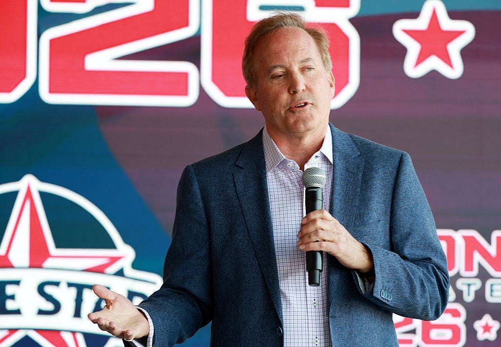 Ken Paxton speaks while holding a microphone in one hand, with a campaign poster in the background.