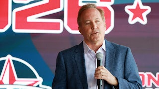 Ken Paxton speaks while holding a microphone in one hand, with a campaign poster in the background.