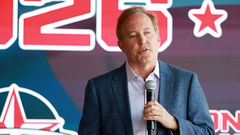 Ken Paxton speaks while holding a microphone in one hand, with a campaign poster in the background.