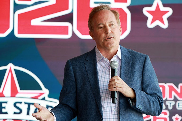 Ken Paxton speaks while holding a microphone in one hand, with a campaign poster in the background.