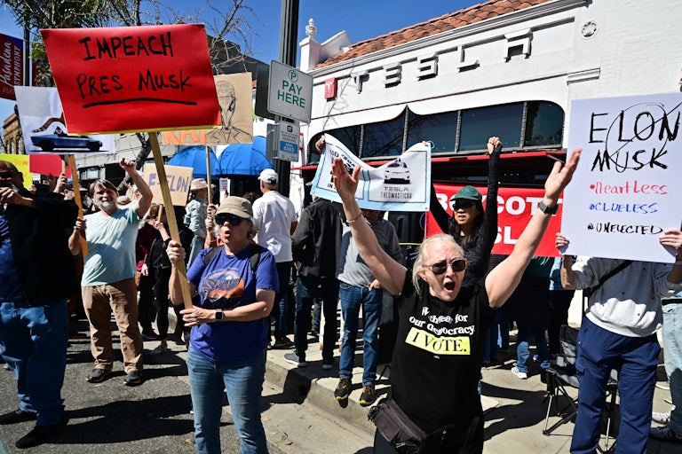 People protest in front of a Tesla dealership. One sign in the foreground reads "Impeach Pres. Musk."