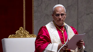 Pope Leo looks to the side while standing in front of a microphone. He holds a stack of papers.