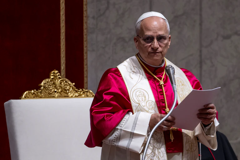Pope Leo looks to the side while standing in front of a microphone. He holds a stack of papers.
