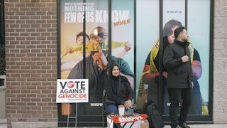 A hijabi woman and a likely Arab man sit/stand near a sign that says "Vote against genocide"