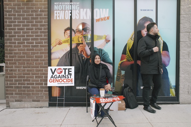 A hijabi woman and a likely Arab man sit/stand near a sign that says "Vote against genocide"
