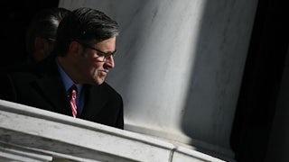 House Speaker Mike Johnson pushes his mouth the side while sitting during a ceremony at Arlington National Cemetery