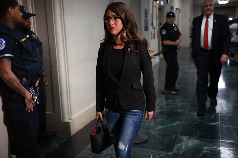 Representative Lauren Boebert walks down a hallway. She is wearing jeans, a black shirt, a black blazer, and has a black purse in her hand. She looks serious and/or distressed. Three security guards are nearby and another man wearing a suit is in the background..