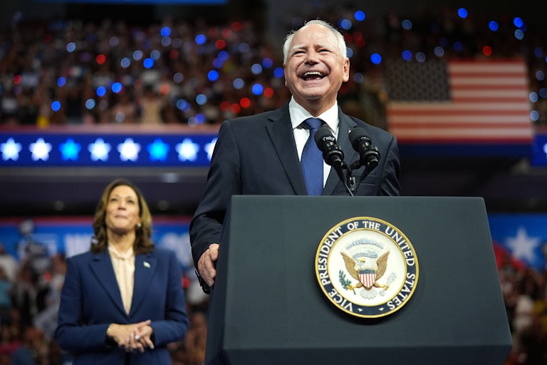 Tim Walz smiles at a podium at a campaign rally. Kamala Harris stands behind him smiling.