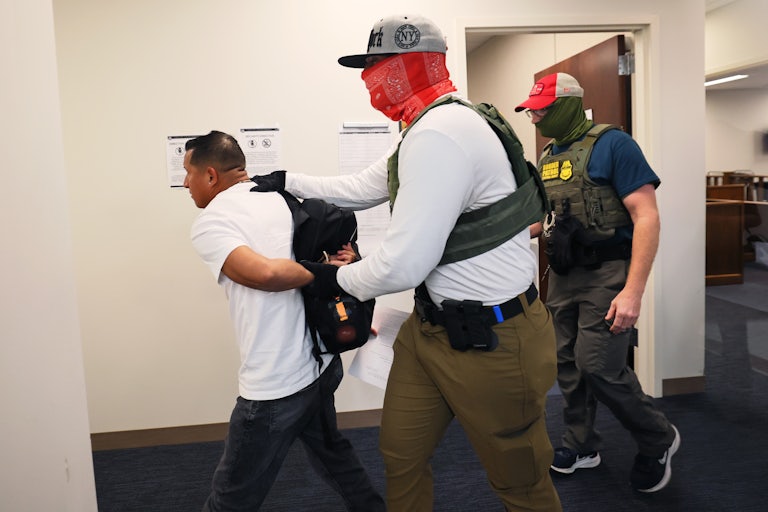 Two agents wearing balaclavas and caps lead a man out of the building with his arms behind his back. One wears a vest that says "Border Patrol."