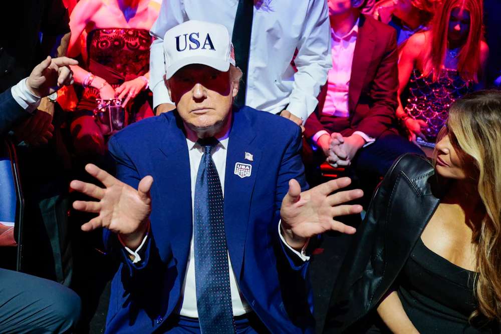 A low energy trump holds out his hands while wearing a white USA hat