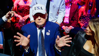 A low energy trump holds out his hands while wearing a white USA hat