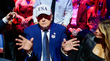 A low energy trump holds out his hands while wearing a white USA hat