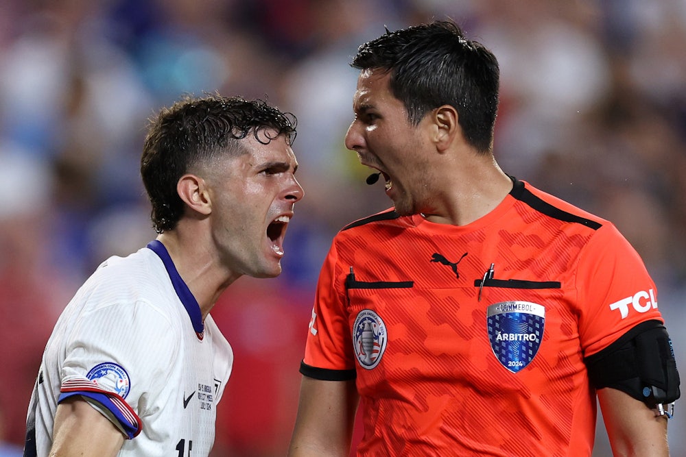 Cristian Pulisic, wearing white, screams in the face of referee Kevin Ortega, wearing an orange official's shirt.