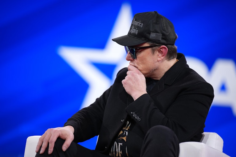 Elon Musk puts his chin in his hand and looks forlorn. He is seated on the CPAC stage and wearing a black MAGA hat, giant black sunglasses, and all black clothes.