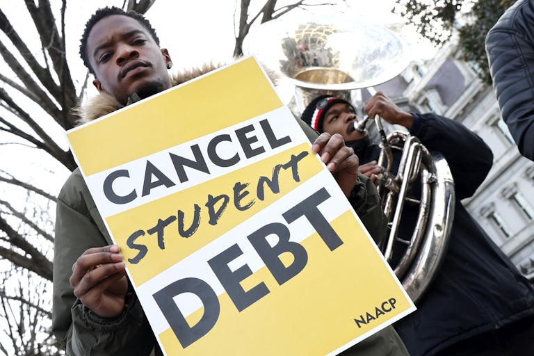 a man holds a sign reading "cancel student debt" while another man plays the tuba
