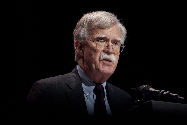 John Bolton wears a suit and speaks at a mic. The background is black.