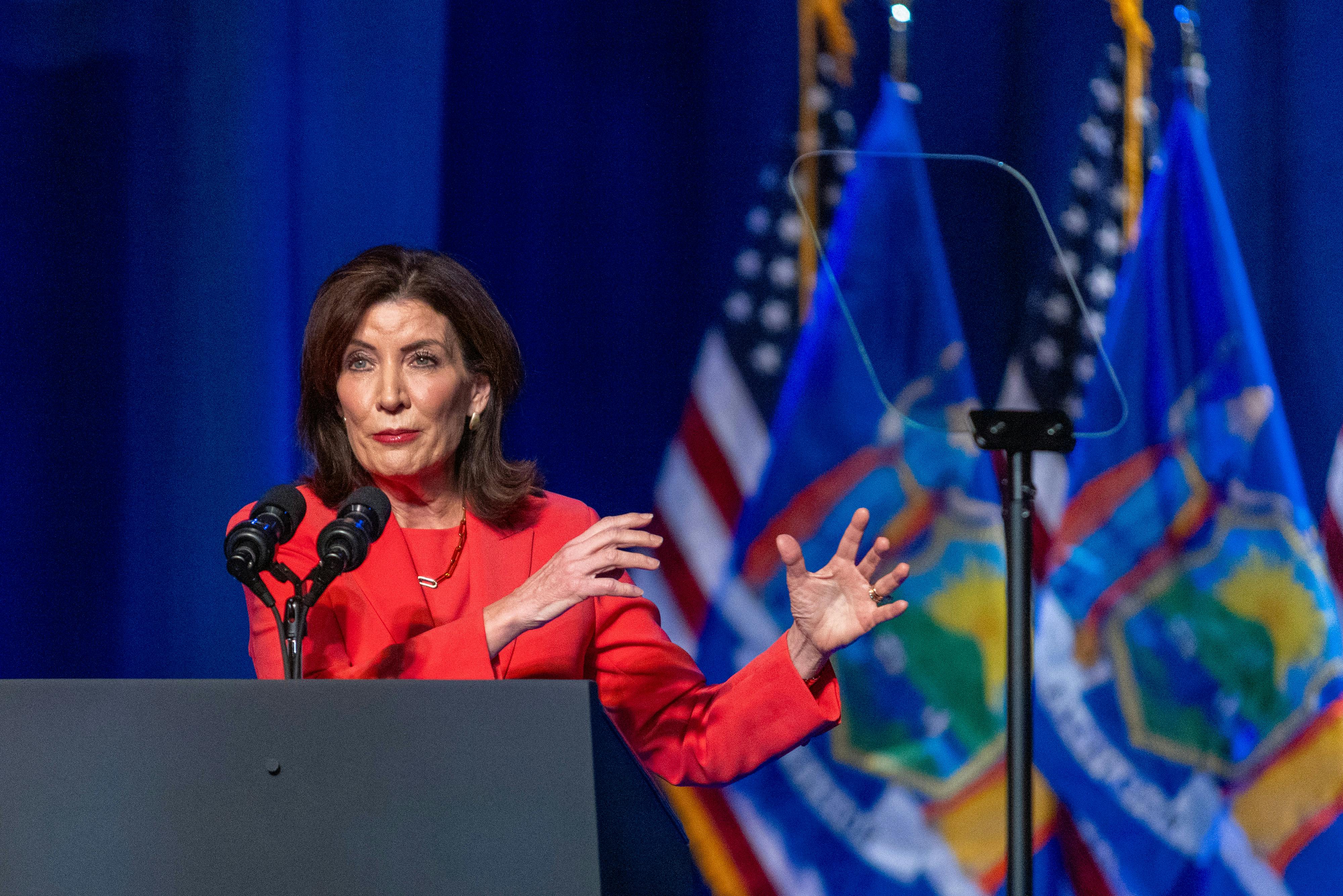 Kathy Hochul, wearing red, gestures to her left while speaking at a podium.