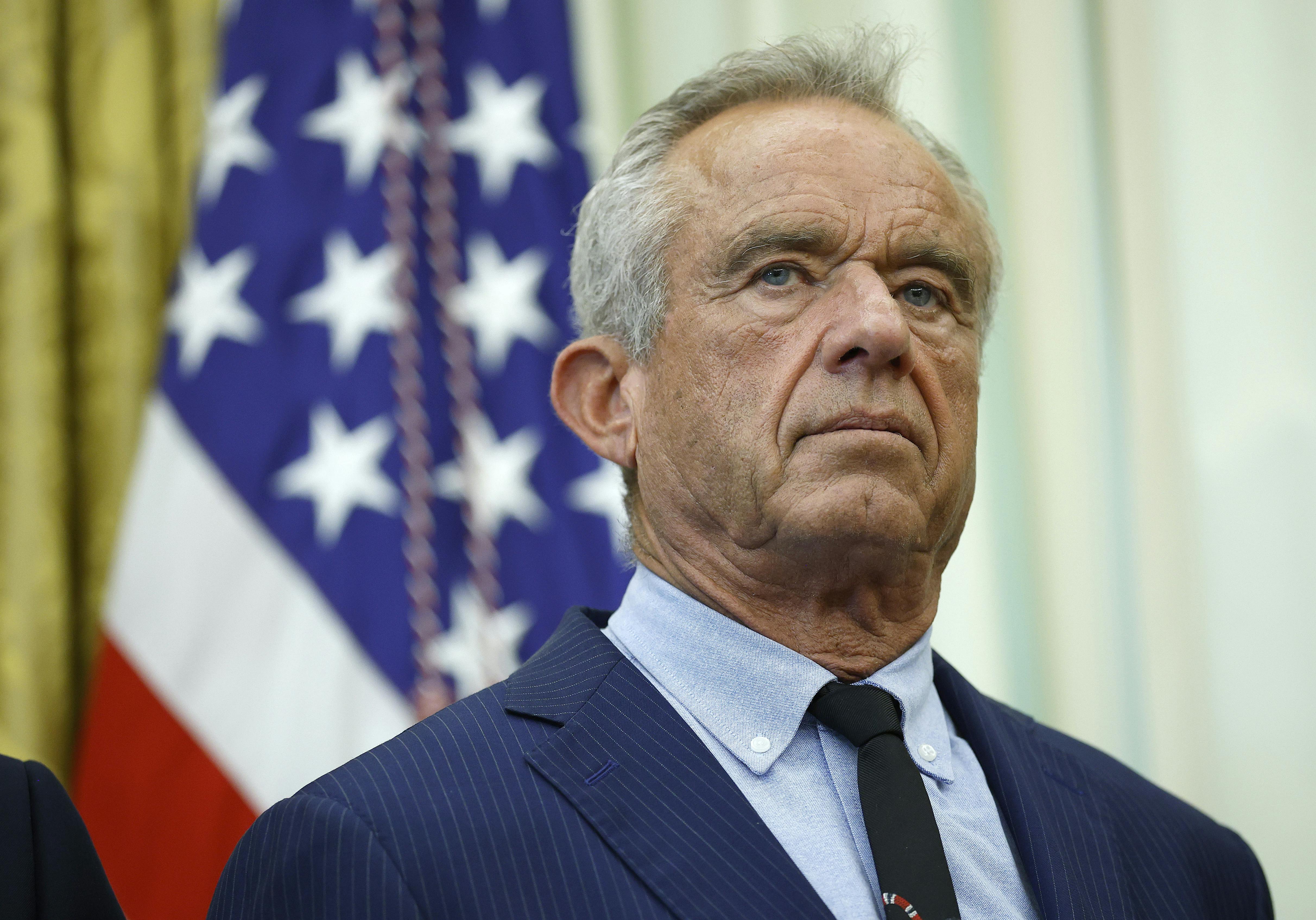 Robert F. Kennedy Jr., wearing a suit and tie, stands in front of American flags at the White House.
