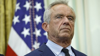 Robert F. Kennedy Jr., wearing a suit and tie, stands in front of American flags at the White House.