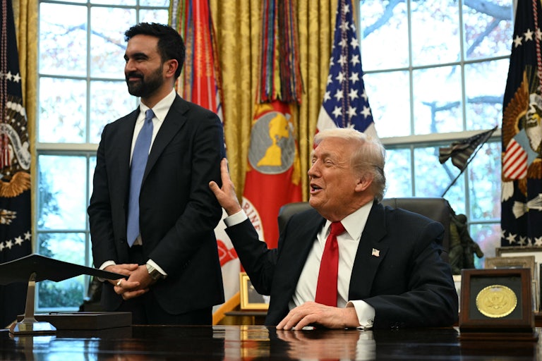 Donald Trump smiles and pats Zohran Mamdani on the arm while sitting at his desk in the Oval Office