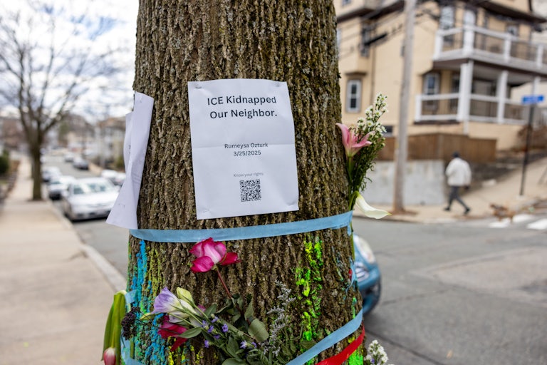 A piece of paper on a tree reads "ICE Kidnapped Our Neighbor. Rumeyza Ozturk 03/25/2025." There is a QR code on the paper, and lots of flowers are also attached to the tree with tape.