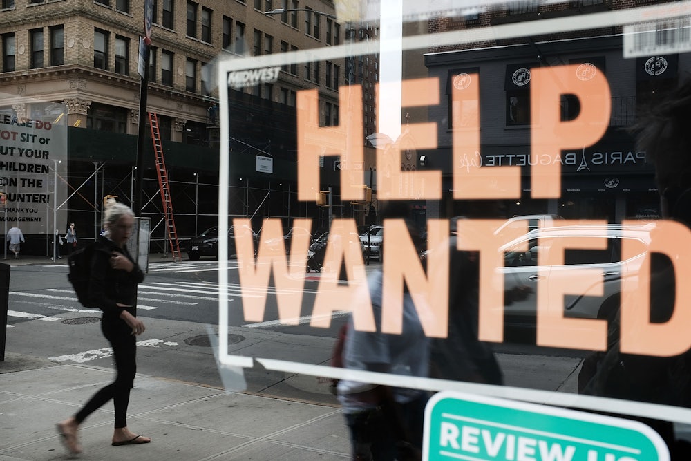 A woman is seen walking down the street in the reflection of a "Help Wanted" sign in a storefront window.