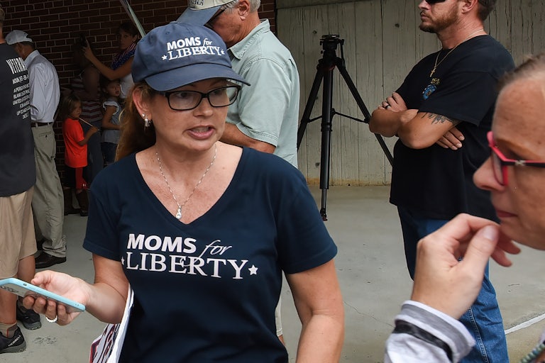 A woman wearing a Moms for Liberty t-shirt and cap hold a phone in her hand and is talking to a man next to her. Others are in the background.