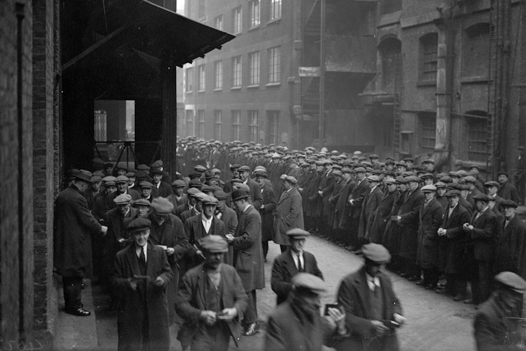 A large quantity of men stand assembled in a dockyard.