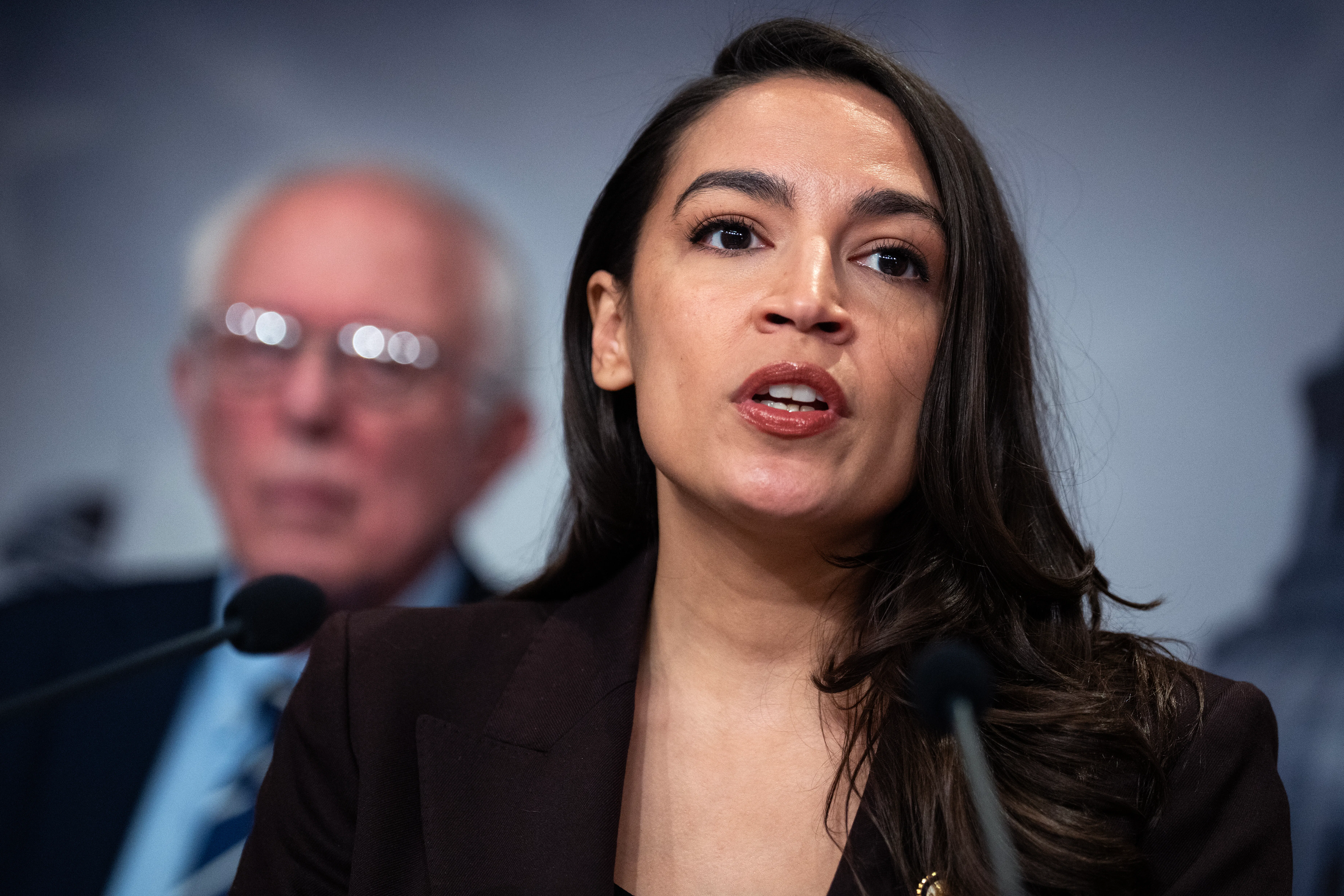 Alexandria Ocasio-Cortez is shown close up, speaking into a microphone, with Bernie Sanders in the background.