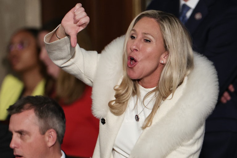 Marjorie Taylor Greene stands up and does a thumbs down gesture while shouting.