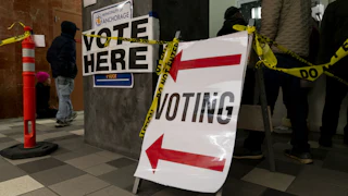 People stand in line to vote. Voting signs and Do Not Enter tape are hung up.