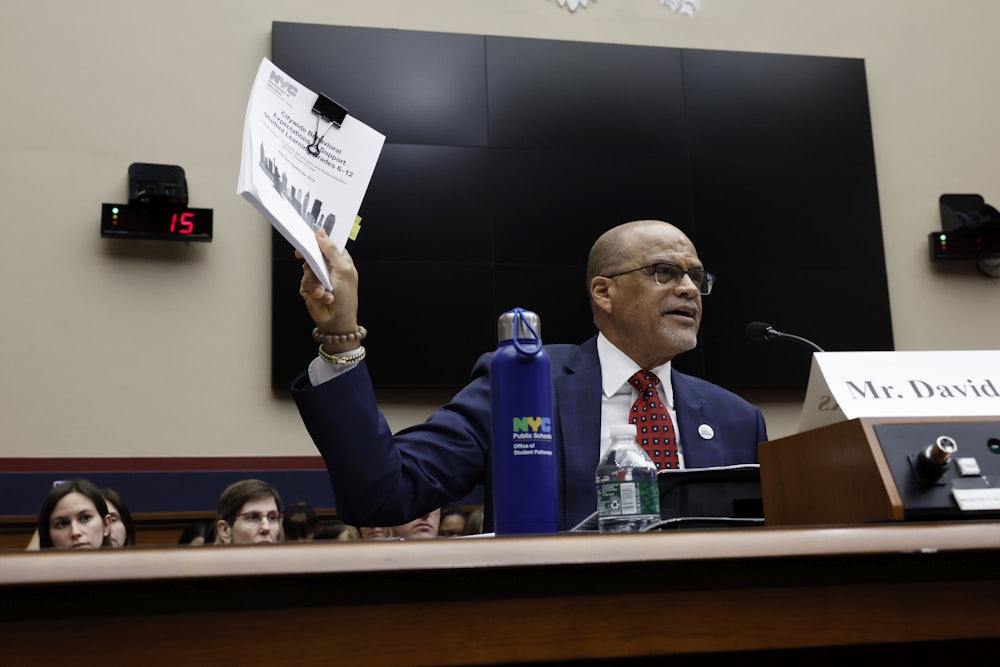 David Banks holds up a stack of papers while sitting at a lectern with people behind him.