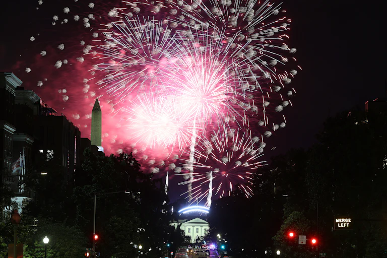 Fireworks are visible in the sky above Washington, D.C., with the Washington Monument and White House visible in the background.