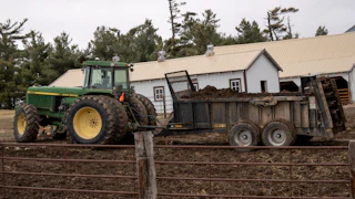 A green tractor pulls a green manure spreader on a dirt field with two buildings behind it and a metal fence in front.
