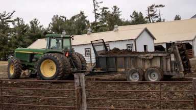 A green tractor pulls a green manure spreader on a dirt field with two buildings behind it and a metal fence in front.