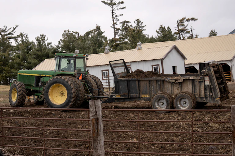 A green tractor pulls a green manure spreader on a dirt field with two buildings behind it and a metal fence in front.