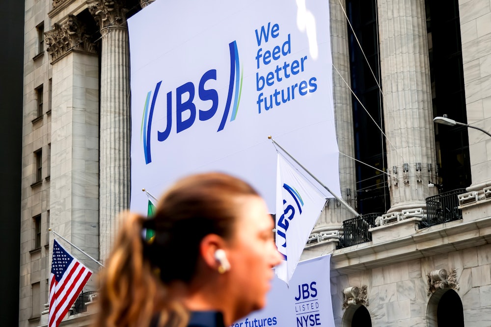 A big sign on the side of a building says "JBS. We feed better futures." A head with a ponytail, out of focus, is seen in the foreground.