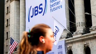 A big sign on the side of a building says "JBS. We feed better futures." A head with a ponytail, out of focus, is seen in the foreground.