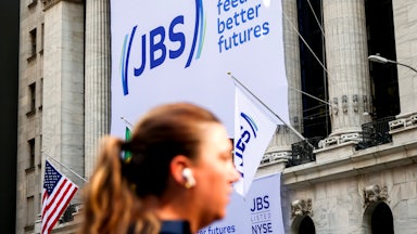 A big sign on the side of a building says "JBS. We feed better futures." A head with a ponytail, out of focus, is seen in the foreground.