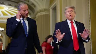 John Thune, left, in a navy suit and dark tie, holds his ear while Donald Trump speaks while wearing a blue suit and red tie.