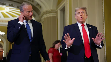 John Thune, left, in a navy suit and dark tie, holds his ear while Donald Trump speaks while wearing a blue suit and red tie.