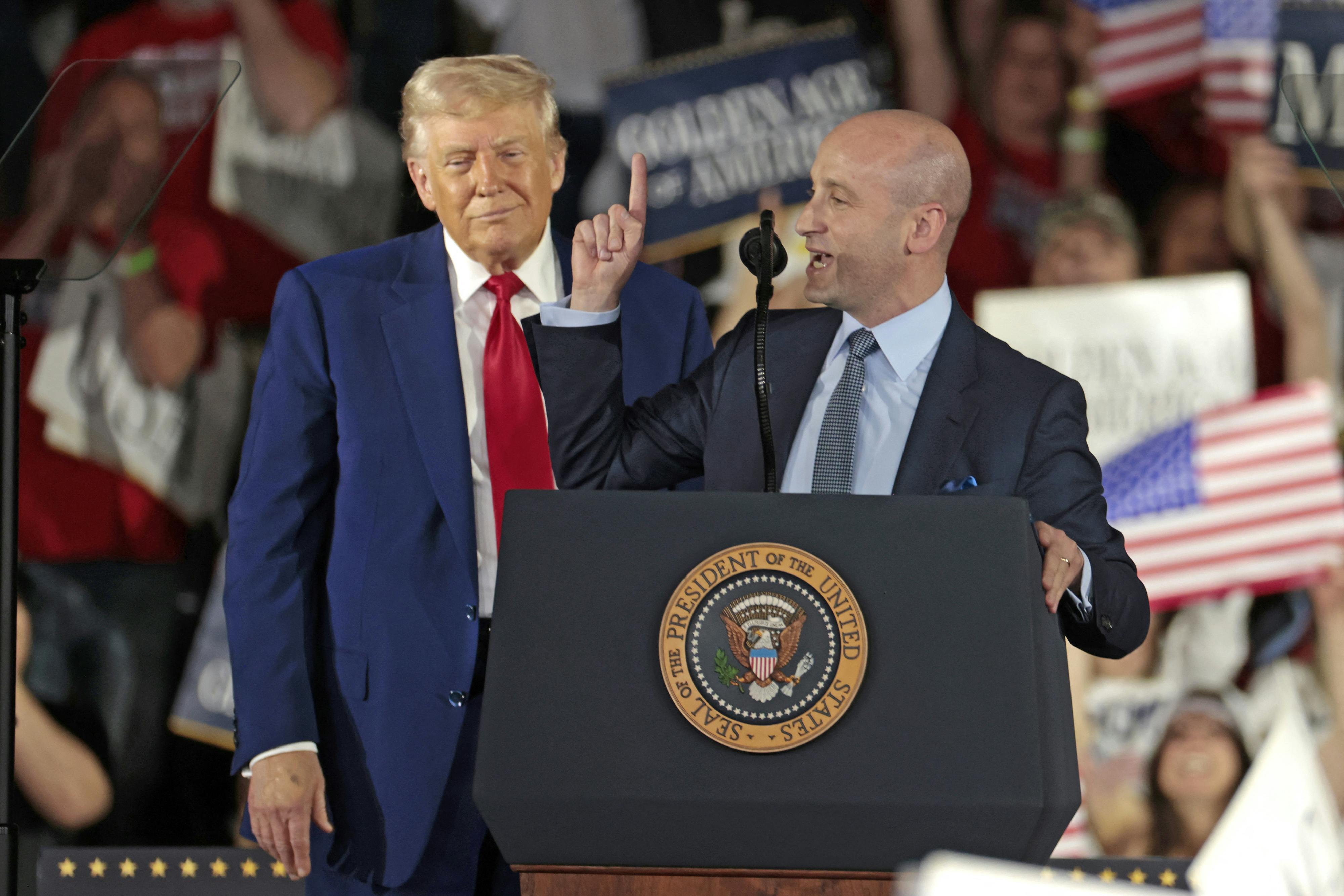 Stephen Miller raises his finger and speaks at a podium while Donald Trump stands behind him