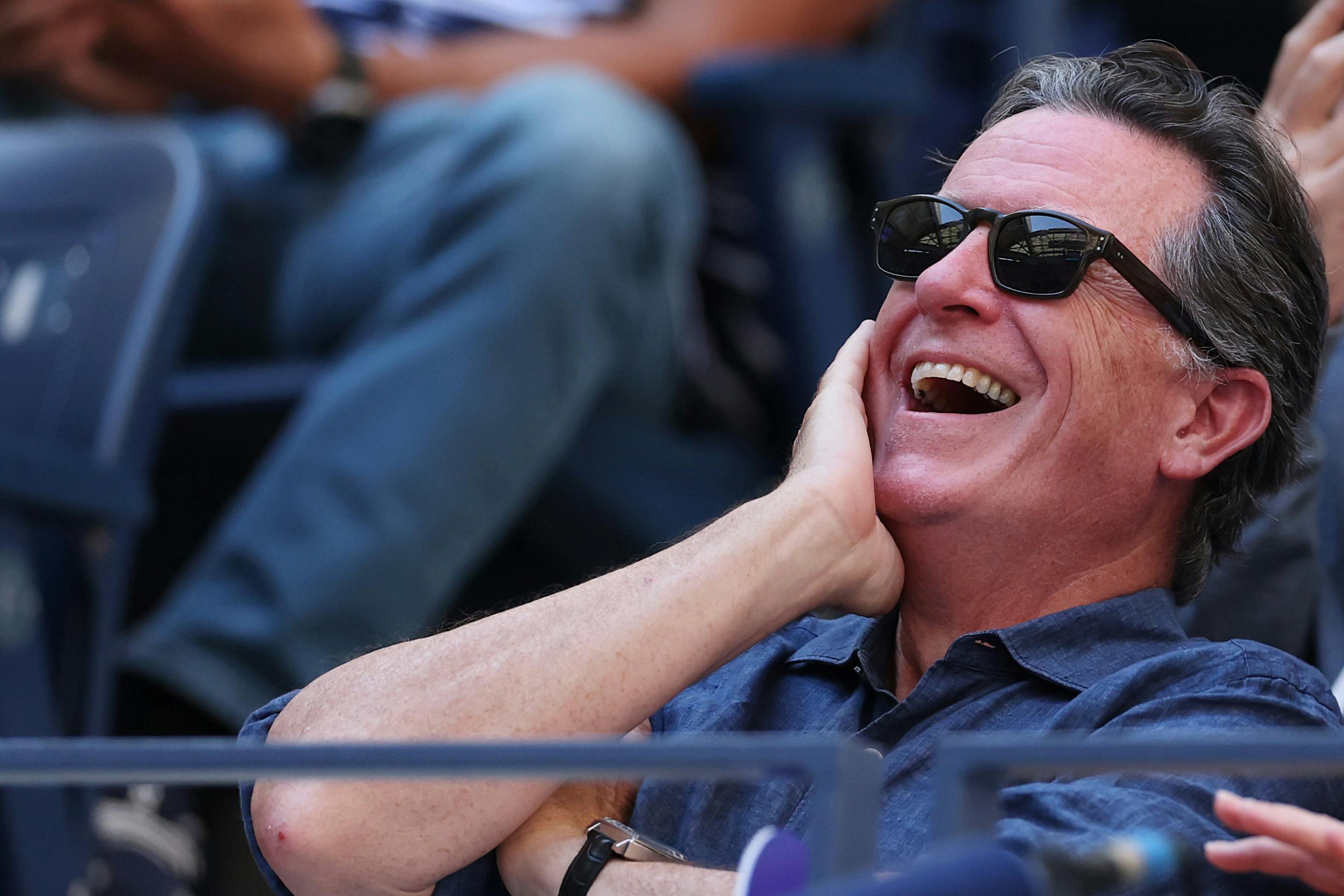 Stephen Colbert laughs while sitting in the stands at a stadium.