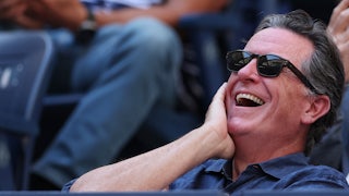 Stephen Colbert laughs while sitting in the stands at a stadium.