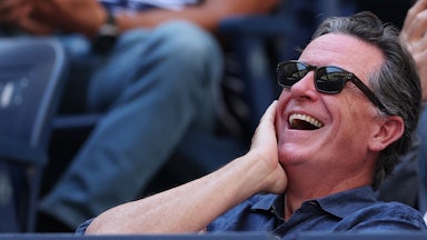 Stephen Colbert laughs while sitting in the stands at a stadium.