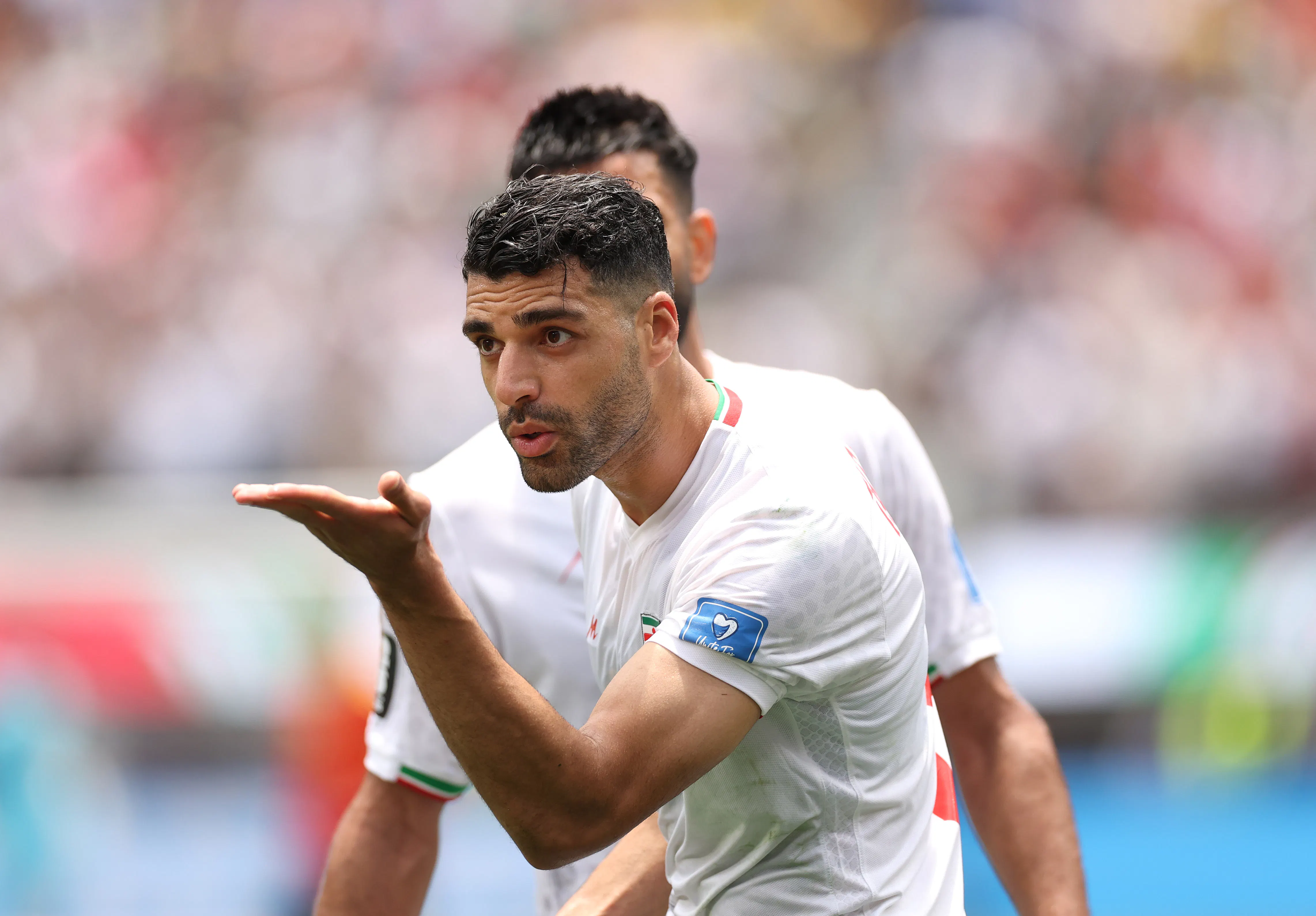 Mehdi Taremi holding his palm out, wearing Iran's white national soccer team jersey, with another obscured player behind him.