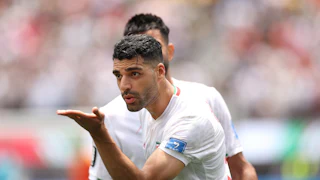 Mehdi Taremi holding his palm out, wearing Iran's white national soccer team jersey, with another obscured player behind him.