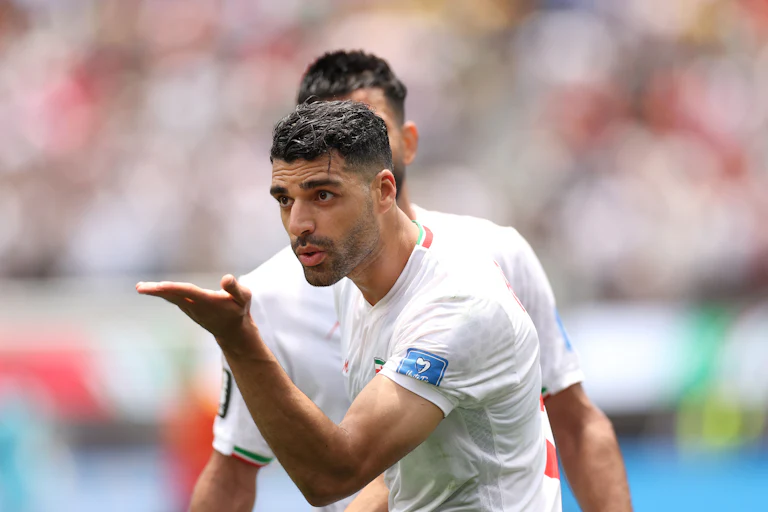 Mehdi Taremi holding his palm out, wearing Iran's white national soccer team jersey, with another obscured player behind him.