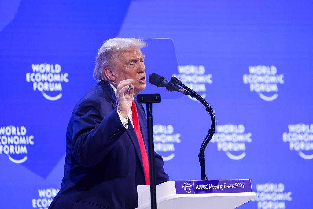 President Trump is seen in profile as he gestures while speaking at a podium labeled "Annual Meeting Davos 2026." Behind him is a purple screen with "World Economic Forum" printed on it.